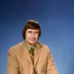 Ralph Klein in a studio portrait from his years as a Calgary television reporter, tan jacket against a blue backdrop