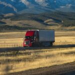A red semi truck crossing golden grassland below rolling hills