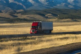 A red semi truck crossing golden grassland below rolling hills