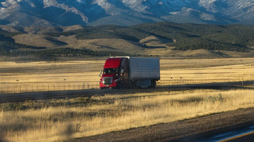 A red semi truck crossing golden grassland below rolling hills