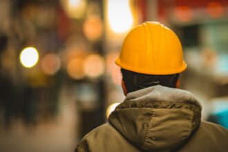 A worker in an orange hard hat and work jacket seen from behind