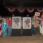 Two framed paintings of a bull rider on a bucking bull on display in an arena, with people standing beside them