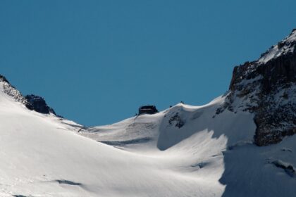 Snow-covered slope rising to a rocky notch between two peaks, with a small hut visible on the skyline of the pass