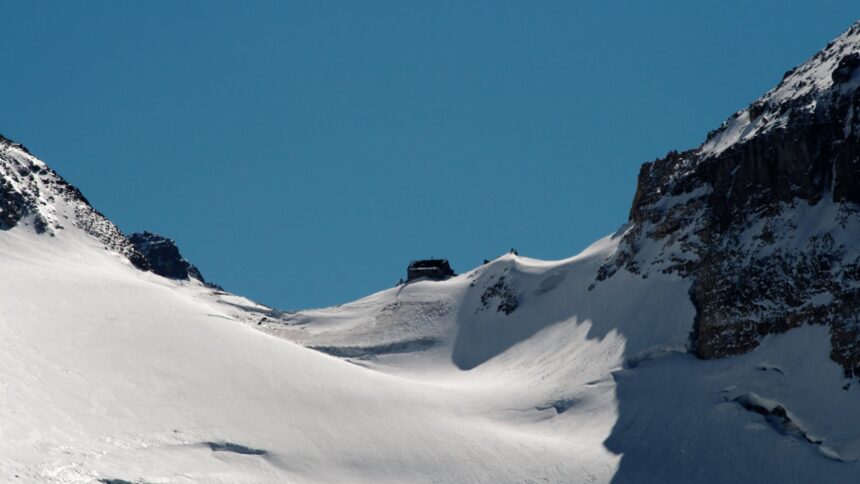 Snow-covered slope rising to a rocky notch between two peaks, with a small hut visible on the skyline of the pass