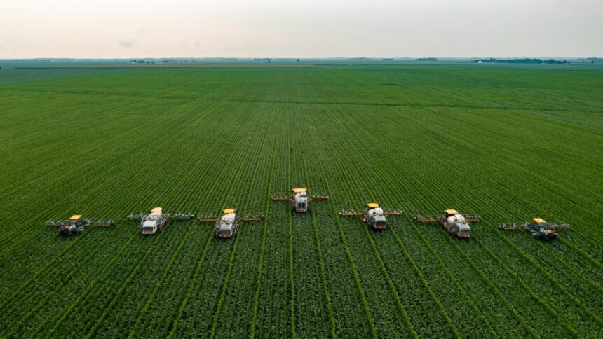 Aerial view of seven agricultural spraying drones flying in a line over a green field
