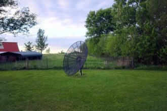 Satellite dish standing on a lawn beside a red barn and trees