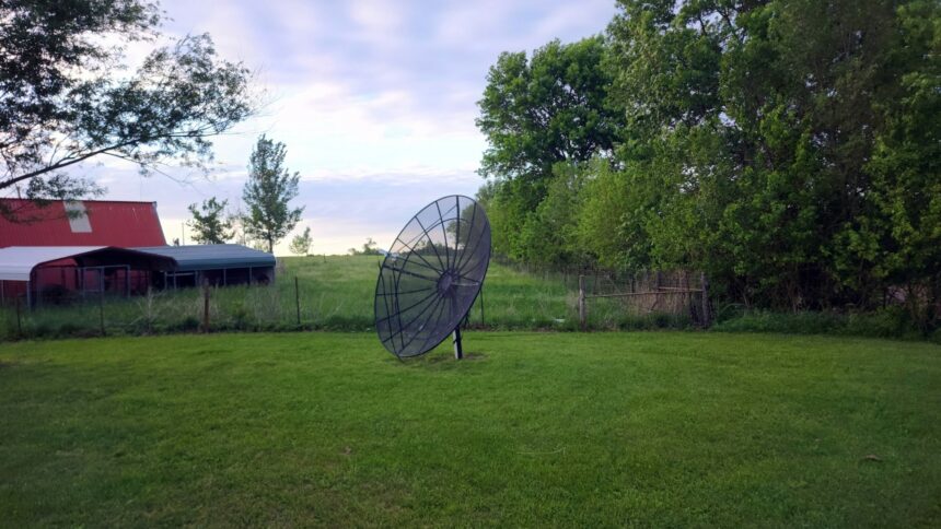Satellite dish standing on a lawn beside a red barn and trees