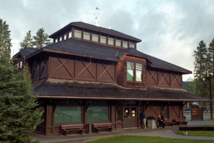 Dark log-faced Banff Park Museum with a tiered shingle roof and a lantern, seen across a lawn with evergreen trees