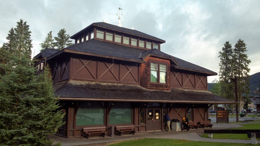 Dark log-faced Banff Park Museum with a tiered shingle roof and a lantern, seen across a lawn with evergreen trees