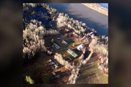 Aerial view of a scatter of small buildings on a riverbank among dry grass, with the river beside them