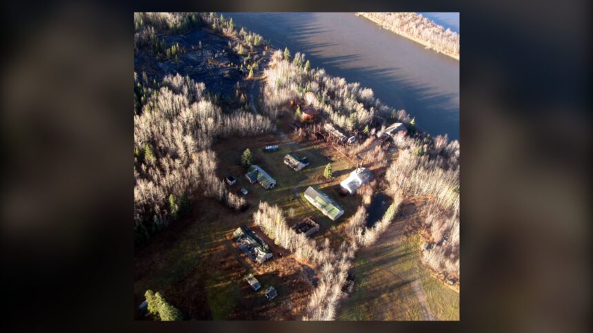 Aerial view of a scatter of small buildings on a riverbank among dry grass, with the river beside them