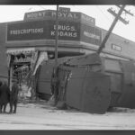 A streetcar lying on its side in the wrecked corner of a brick drugstore building, with onlookers and a leaning utility pole, in a black and white photograph