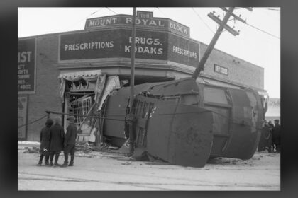 A streetcar lying on its side in the wrecked corner of a brick drugstore building, with onlookers and a leaning utility pole, in a black and white photograph
