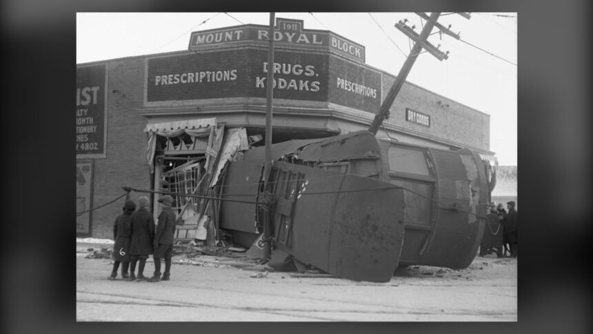 A streetcar lying on its side in the wrecked corner of a brick drugstore building, with onlookers and a leaning utility pole, in a black and white photograph