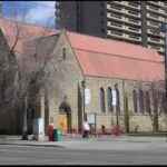 Rusticated sandstone Cathedral Church of the Redeemer with a steep red metal roof, seen from a street corner in Calgary with high-rise towers behind