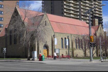 Rusticated sandstone Cathedral Church of the Redeemer with a steep red metal roof, seen from a street corner in Calgary with high-rise towers behind