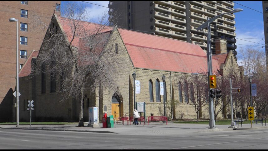 Rusticated sandstone Cathedral Church of the Redeemer with a steep red metal roof, seen from a street corner in Calgary with high-rise towers behind