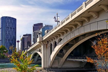 A concrete arch bridge crossing a river beside Calgary office towers, with autumn-coloured trees on the bank