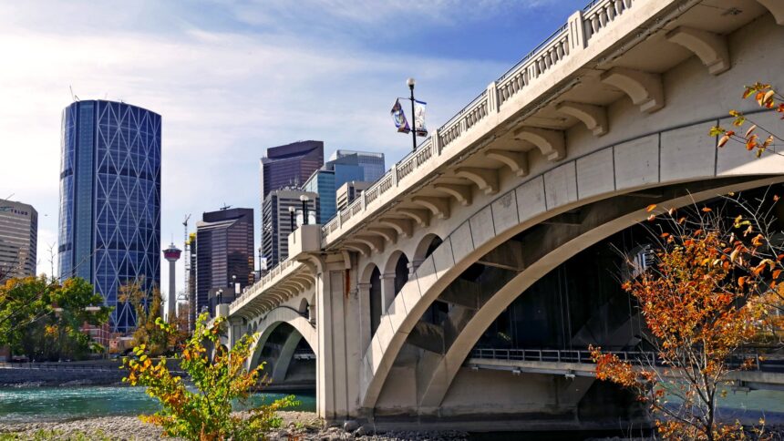 A concrete arch bridge crossing a river beside Calgary office towers, with autumn-coloured trees on the bank