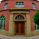 Red brick building with two arched windows in green frames either side of a pair of wooden doors in a sandstone surround, with a lamp post and hanging flower baskets on the sidewalk