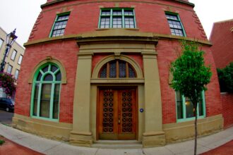 Red brick building with two arched windows in green frames either side of a pair of wooden doors in a sandstone surround, with a lamp post and hanging flower baskets on the sidewalk