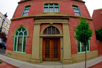 Red brick building with two arched windows in green frames either side of a pair of wooden doors in a sandstone surround, with a lamp post and hanging flower baskets on the sidewalk