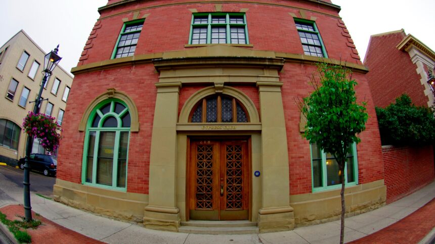 Red brick building with two arched windows in green frames either side of a pair of wooden doors in a sandstone surround, with a lamp post and hanging flower baskets on the sidewalk