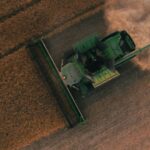 Green combine harvester cutting a wheat field, seen from above, with dust trailing behind