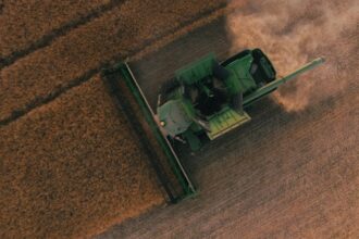 Green combine harvester cutting a wheat field, seen from above, with dust trailing behind