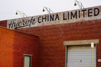 Red brick wall with the words Hycroft China Limited lettered across the top, three green-shaded lamps on curved arms above the sign and a white roll-up door below
