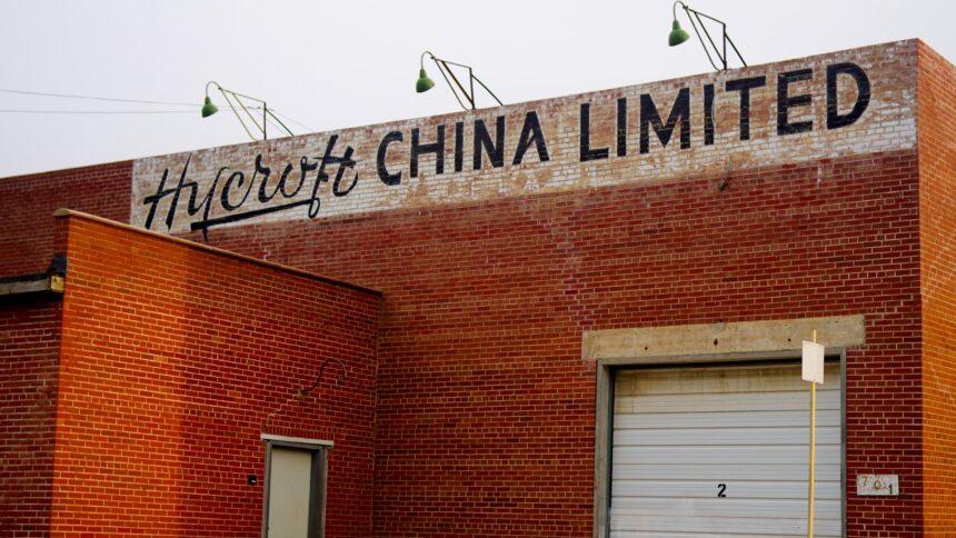 Red brick wall with the words Hycroft China Limited lettered across the top, three green-shaded lamps on curved arms above the sign and a white roll-up door below