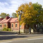 Long one-storey red brick building with a pitched red roof, a tall brick chimney and a brick cornice, seen from across a street in autumn with yellow trees in front