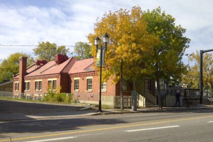 Long one-storey red brick building with a pitched red roof, a tall brick chimney and a brick cornice, seen from across a street in autumn with yellow trees in front