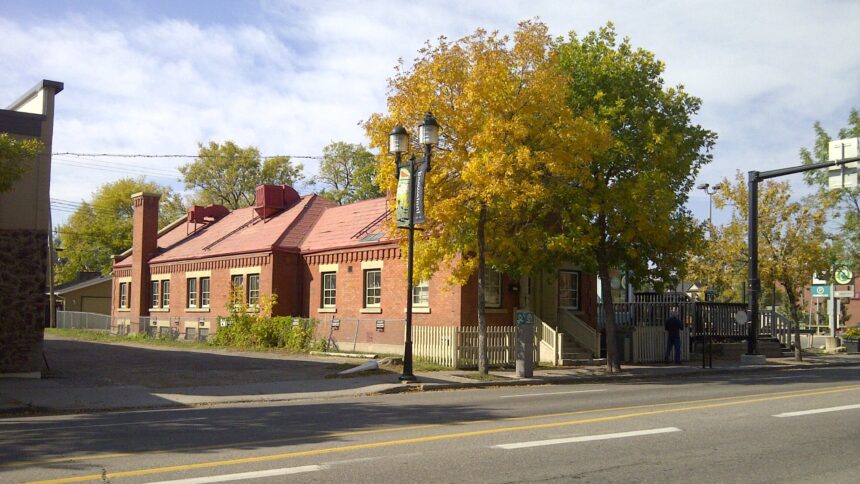 Long one-storey red brick building with a pitched red roof, a tall brick chimney and a brick cornice, seen from across a street in autumn with yellow trees in front