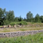 A low dry-laid stone retaining wall runs through grass in the foreground, with a ruined stone gable showing brick patches and scattered stone remnants beyond, under trees and a clear sky
