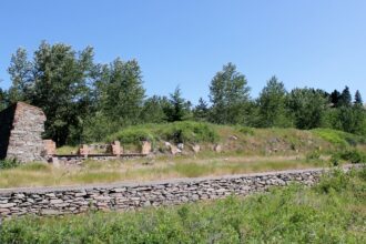 A low dry-laid stone retaining wall runs through grass in the foreground, with a ruined stone gable showing brick patches and scattered stone remnants beyond, under trees and a clear sky