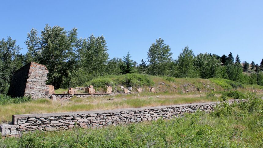 A low dry-laid stone retaining wall runs through grass in the foreground, with a ruined stone gable showing brick patches and scattered stone remnants beyond, under trees and a clear sky