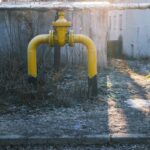 Yellow natural gas riser pipe with a shut-off valve standing in frosted grass beside a concrete wall, low autumn sun casting long shadows
