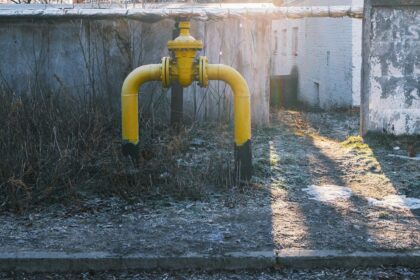 Yellow natural gas riser pipe with a shut-off valve standing in frosted grass beside a concrete wall, low autumn sun casting long shadows