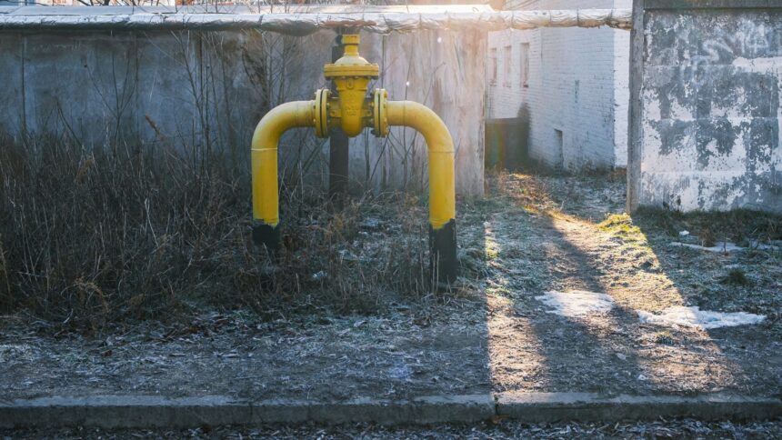 Yellow natural gas riser pipe with a shut-off valve standing in frosted grass beside a concrete wall, low autumn sun casting long shadows