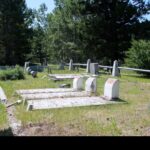Grave markers and flat concrete slabs in the western plot of Old Hillcrest Cemetery, with an angled interpretive panel in the foreground, a fence and forested hills behind