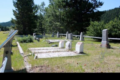Grave markers and flat concrete slabs in the western plot of Old Hillcrest Cemetery, with an angled interpretive panel in the foreground, a fence and forested hills behind