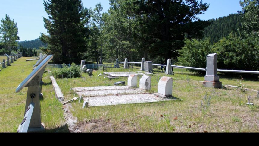 Grave markers and flat concrete slabs in the western plot of Old Hillcrest Cemetery, with an angled interpretive panel in the foreground, a fence and forested hills behind