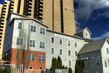 Grey-sided Ritchie Mill with a brick lower storey and the name painted on its wall, beside high-rise towers in Edmonton