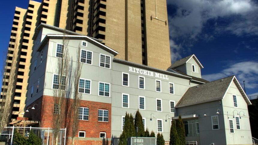 Grey-sided Ritchie Mill with a brick lower storey and the name painted on its wall, beside high-rise towers in Edmonton