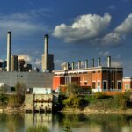 Brick power plant with a row of small roof stacks, a grey block and taller stacks beyond, across a river with downtown towers behind