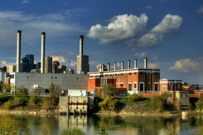 Brick power plant with a row of small roof stacks, a grey block and taller stacks beyond, across a river with downtown towers behind