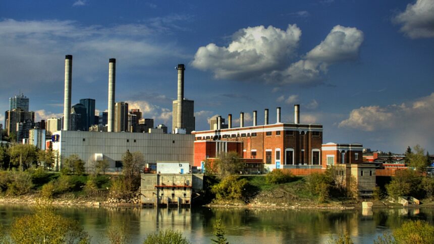 Brick power plant with a row of small roof stacks, a grey block and taller stacks beyond, across a river with downtown towers behind