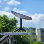 Flat satellite internet dish mounted on a pole against a blue sky with clouds and green trees