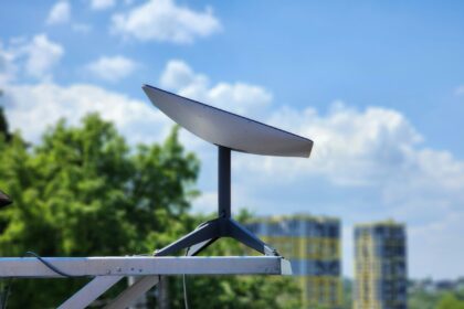 Flat satellite internet dish mounted on a pole against a blue sky with clouds and green trees